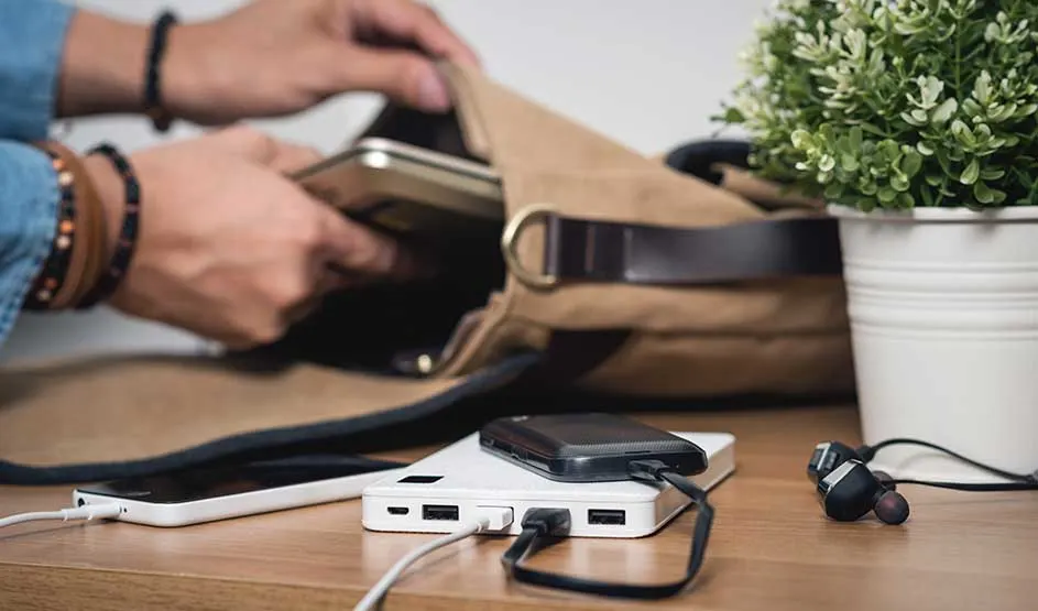 Sur une table en bois reposent batteries externes, câbles et écouteurs, tandis qu’une personne range son smartphone dans son sac.