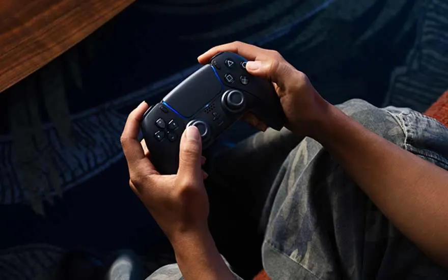 Un joueur tient une manette PlayStation 4 noire avec des accents bleus dans ses mains, assis sur un canapé avec une table en bois et un éclairage bleu ambiant en arrière-plan.