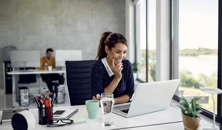 Une femme travaille sur un ordinateur portable à un bureau blanc, avec une tasse et un verre d'eau à proximité. Une fenêtre est visible en arrière-plan.