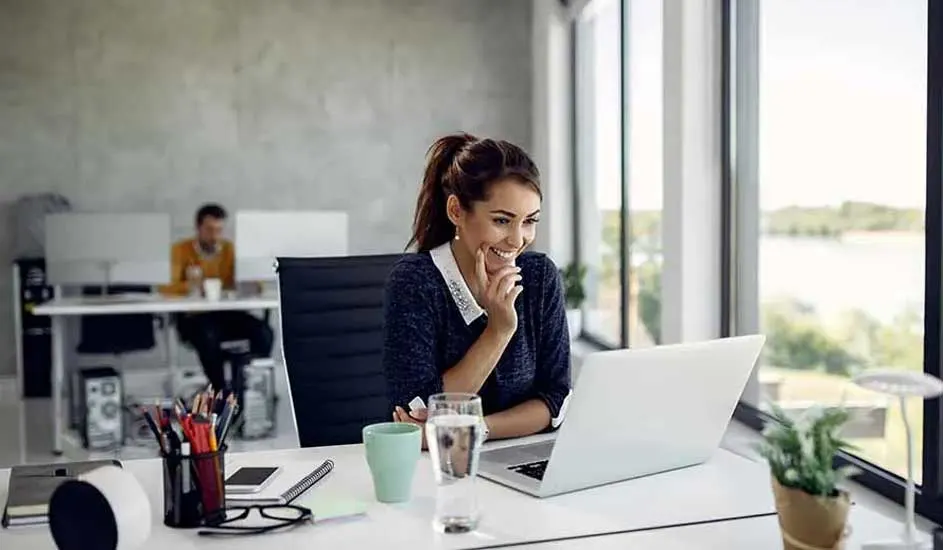 Une femme travaille sur un ordinateur portable à un bureau blanc, avec une tasse et un verre d'eau à proximité. Une fenêtre est visible en arrière-plan.