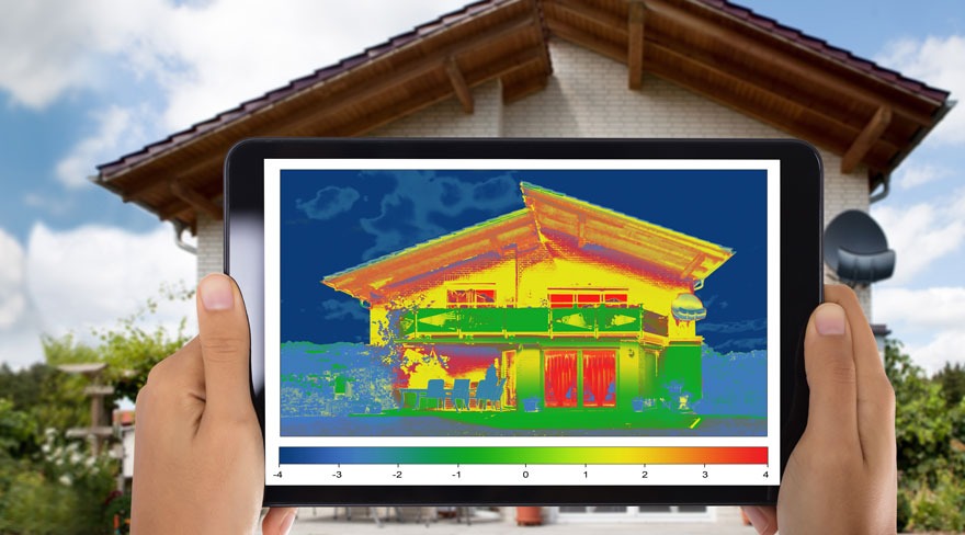 Une tablette affiche l'imagerie thermique d'une maison, avec un toit brun et un extérieur beige, sous un ciel bleu nuageux.