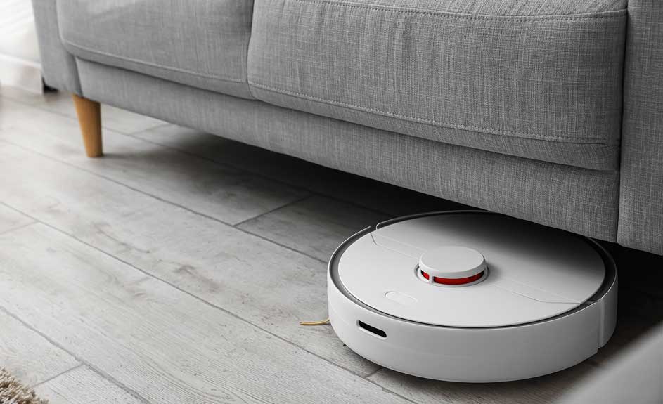 Een witte robotstofzuiger staat onder een grijze zetel op een houten vloer.