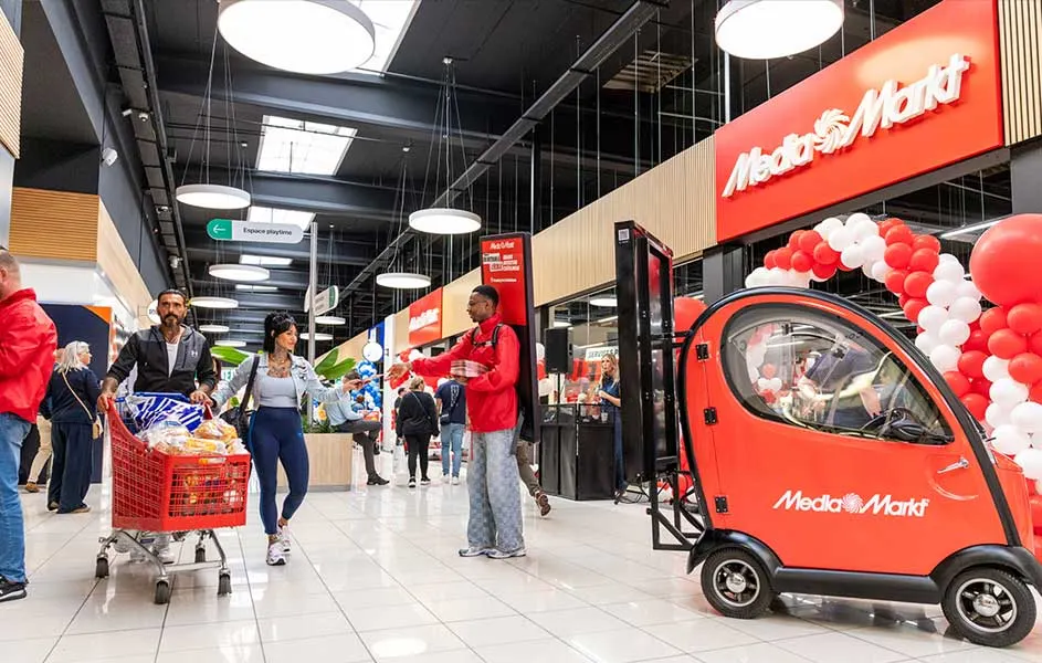 In een ruime MediaMarkt lopen klanten langs schappen en winkelwagens. Een rood promotiewagentje staat naast een boog van rode en witte ballonnen.