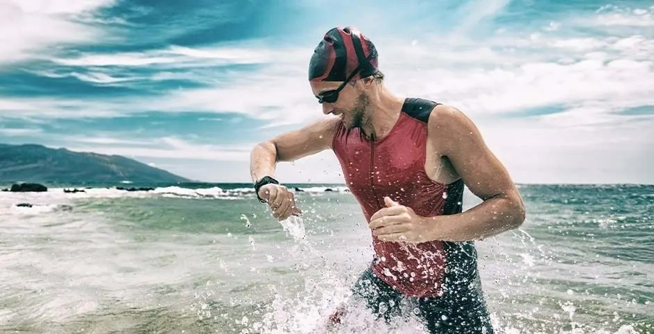 Een atleet in wetsuit en badmuts rent uit zee, water spat op, de lucht is blauw en bergen zijn zichtbaar op de achtergrond.