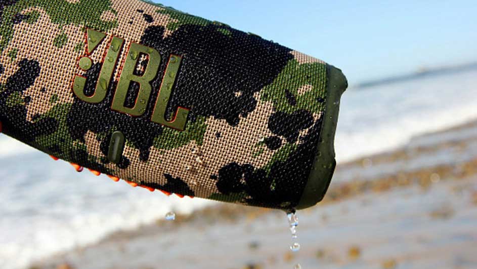 Een camouflage JBL-speaker met waterdruppels tegen een achtergrond van zand en vage blauwe lucht.