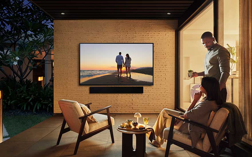 Een gezellig terras in de avond, een vrouw zit ontspannen, een man maakt drankjes klaar, tv toont een koppel aan het strand.