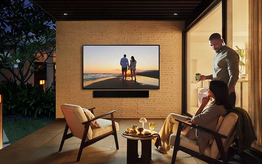 Terrasse chaleureuse en soirée, une femme détendue, un homme prépare des cocktails, téléviseur affiche un couple sur la plage.