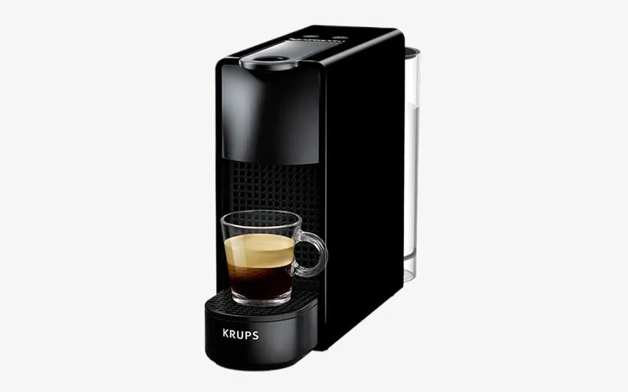 A slim black Krups coffee machine prepares an espresso in a glass cup, with a transparent water tank positioned at the back.