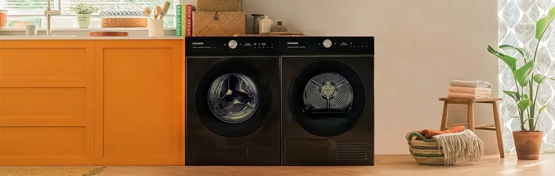 A laundry room features a black washer and dryer set next to orange cabinets. Nearby, a stool holds folded towels and a potted plant.