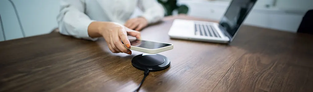 Téléphone sur un chargeur sans fil noir, posé sur une table en bois brun, avec un ordinateur portable en arrière-plan.