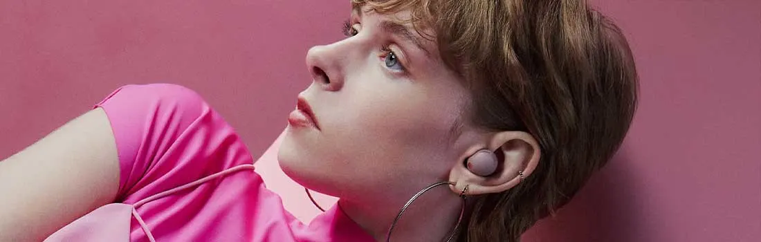 Persoon in felroze top ligt achterover tegen een roze achtergrond, met stijlvolle draadloze oordopjes en grote ronde oorbellen.