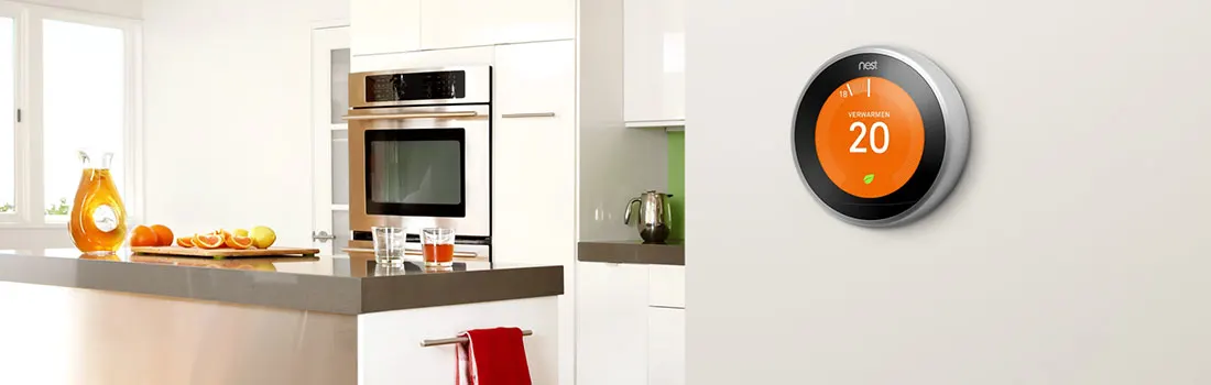 Un thermostat Nest rond affiche 20 degrés sur un mur blanc. Une cuisine moderne est visible en arrière-plan.