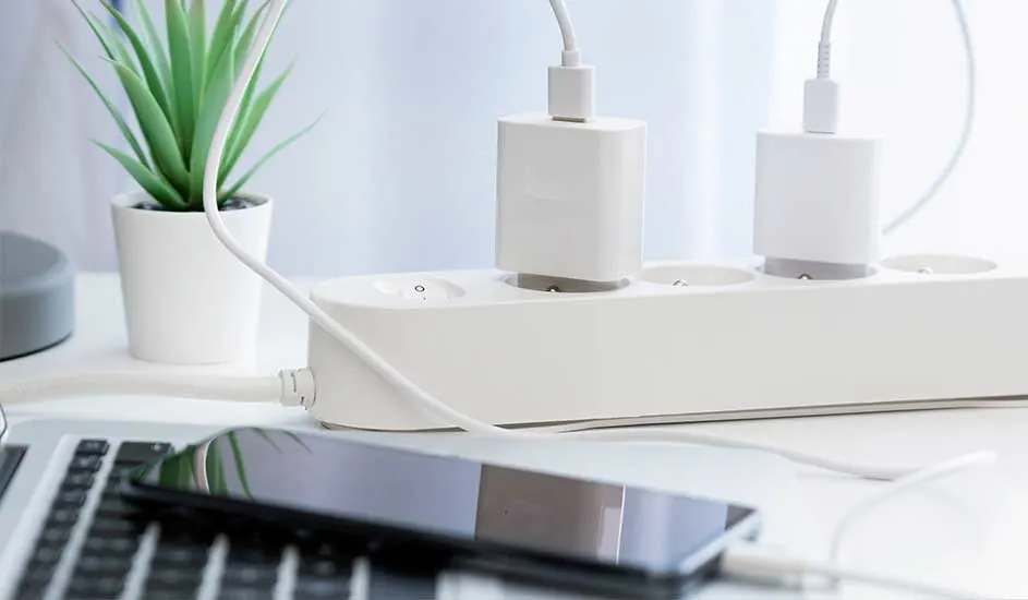 Een wit stekkerblok met twee laders en snoeren. Een laptop, telefoon en plant staan op een witte tafel.