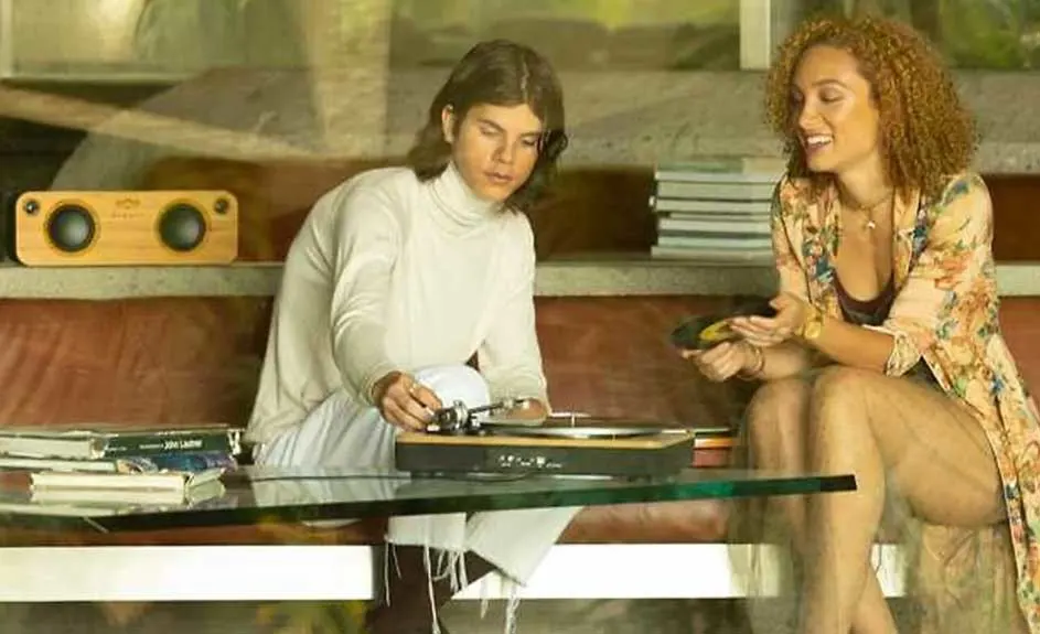 Deux personnes près d'une table en verre. L'une manipule une platine disque, l'autre tient un disque vinyle. Haut-parleur en bois visible.