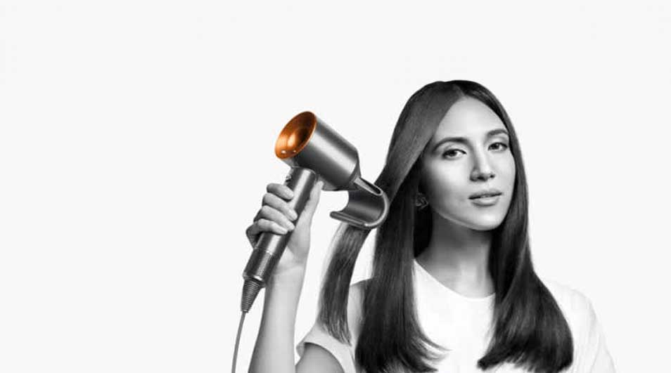 Une main tient un sèche-cheveux gris et orange près de longs cheveux bruns. Le fond est blanc.