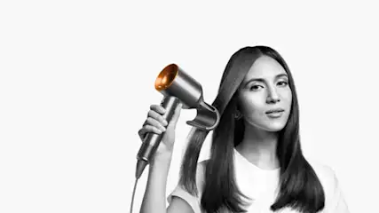 Une main tient un sèche-cheveux gris et orange près de longs cheveux bruns. Le fond est blanc.