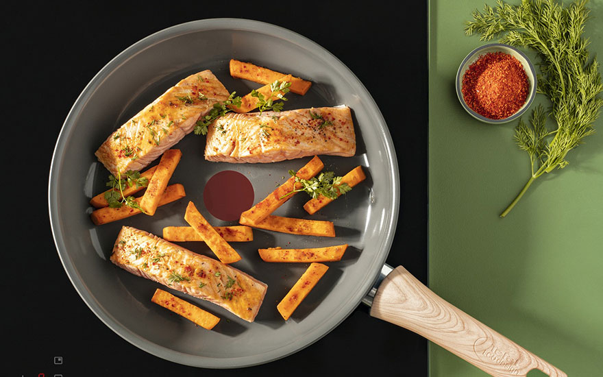 Een grijze pan met zalm en zoete aardappelfrietjes staat op een zwarte kookplaat. Een kom met rood poeder en dille staan op een groene achtergrond.