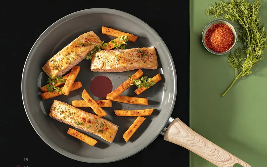 Een grijze pan met zalm en zoete aardappelfrietjes staat op een zwarte kookplaat. Een kom met rood poeder en dille staan op een groene achtergrond.