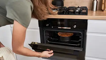 Une cuisine avec un four noir ouvert, révélant un plat rond. Une cuisinière noire est visible sur un comptoir en bois.