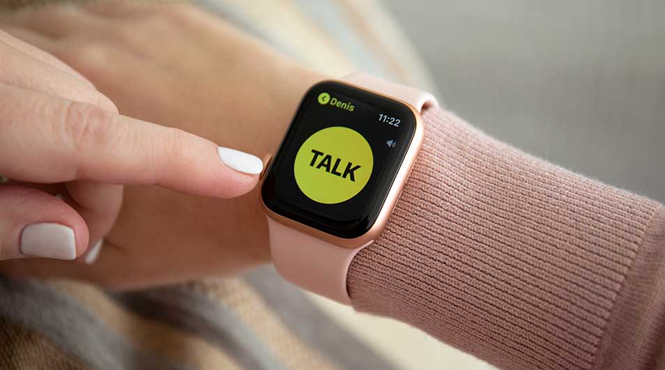 Une personne touche une montre intelligente avec un bracelet rose. L'écran affiche "TALK" en jaune.