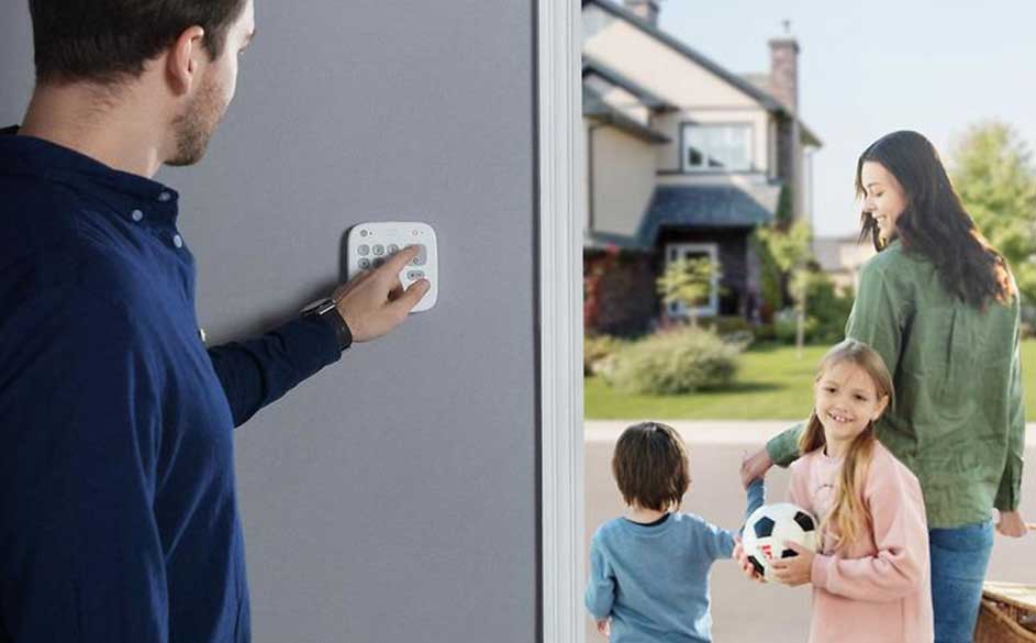 Un homme active un système d'alarme mural. Dehors, une maison et des enfants avec un ballon de foot sur un trottoir.