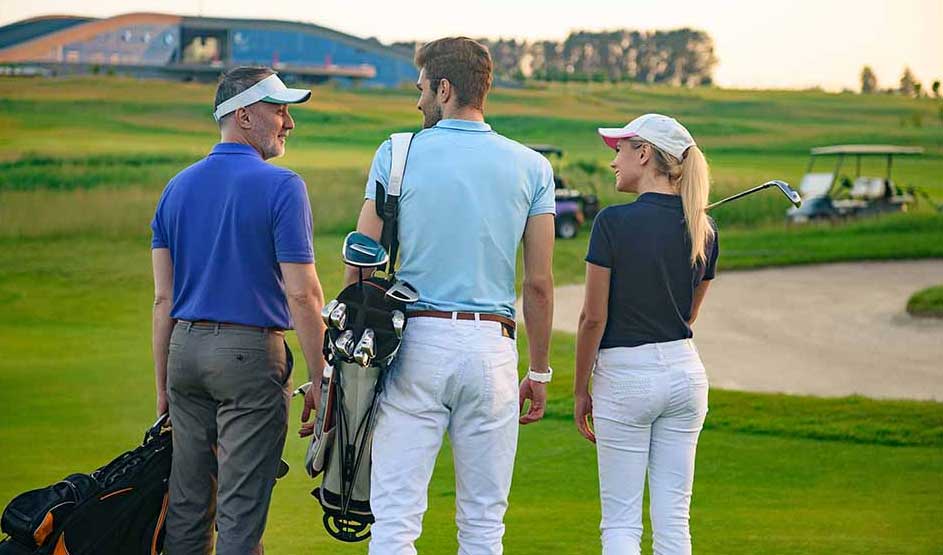 Trois golfeurs marchent côte à côte sur le fairway, sacs à l’épaule, discutant joyeusement dans la lumière douce du soir.