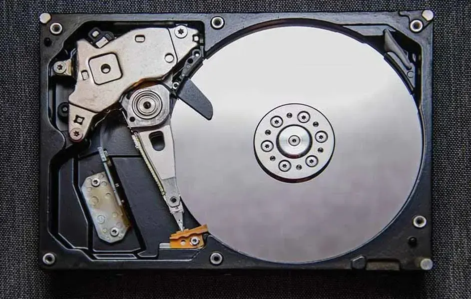 Un disque dur ouvert montre un plateau argenté et des composants mécaniques sur un fond gris texturé.