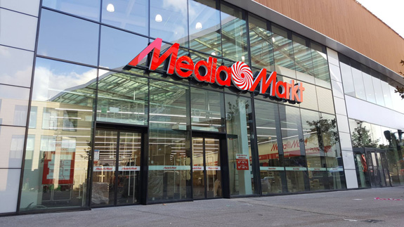 Façade vitrée d’un grand magasin MediaMarkt, logo rouge vif au-dessus des portes coulissantes, intérieur lumineux rempli d’électronique.
