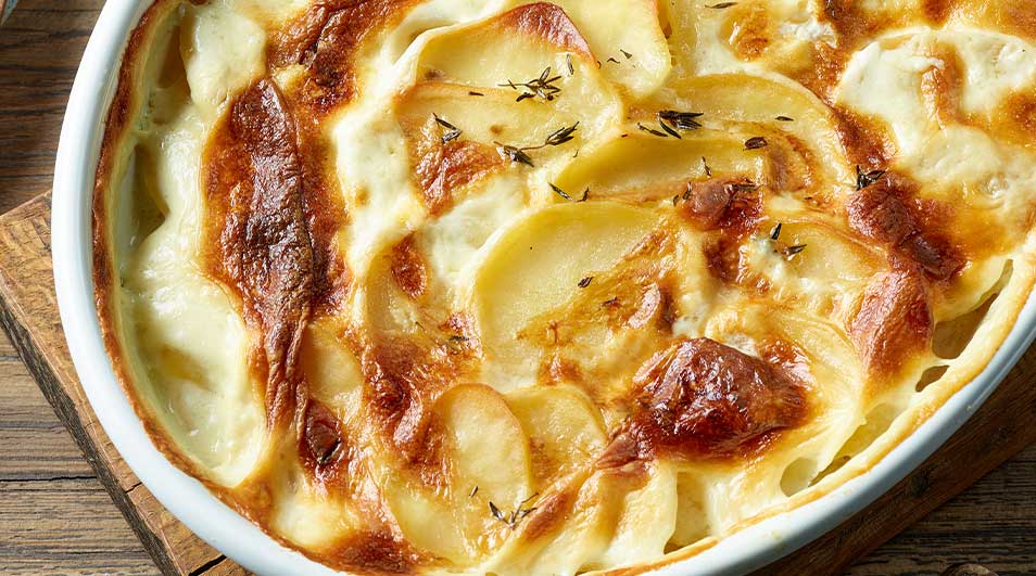 Gratin dauphinois dans un plat blanc ovale, sur une planche en bois. Les pommes de terre sont dorées et parsemées de thym.