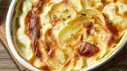 Gratin dauphinois dans un plat blanc ovale, sur une planche en bois. Les pommes de terre sont dorées et parsemées de thym.