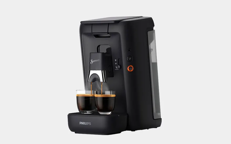 A silver and black Nespresso machine with a glass cup of coffee sits on a grey surface, along with a purple box and coffee capsules.