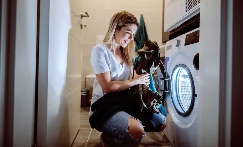 In een badkamer staat een witte wasmachine met een stapel kleren ervoor. Tegels bedekken de vloer en een handdoek hangt aan de muur.