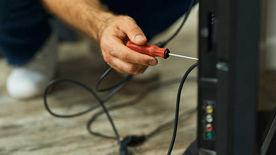 A red screwdriver is used on the back of a black TV, with black cables connected to colored ports. The floor is light brown.
