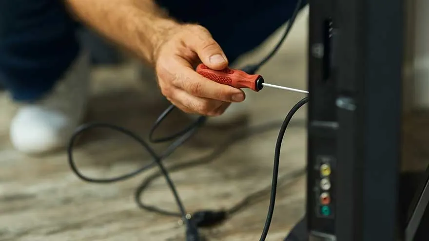 A red screwdriver is used on the back of a black TV, with black cables connected to colored ports. The floor is light brown.