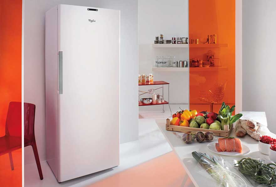 Frigo blanc dans une cuisine avec fruits, légumes et herbes sur une table blanche. Étagères orange en arrière-plan.