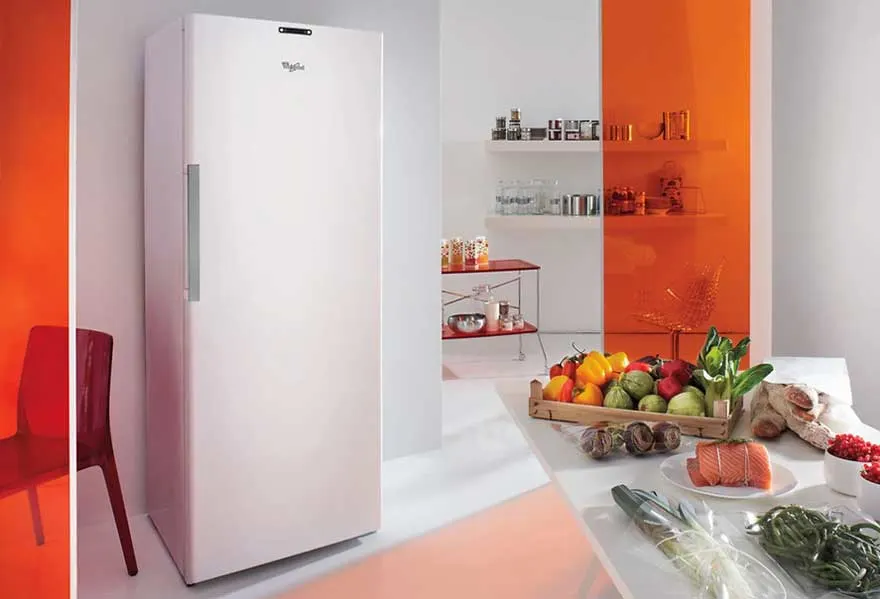 Frigo blanc dans une cuisine avec fruits, légumes et herbes sur une table blanche. Étagères orange en arrière-plan.