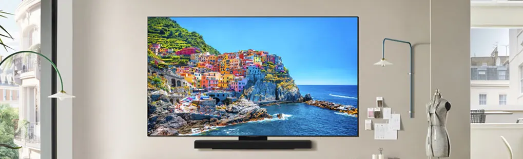 Een flatscreen-tv toont een kustplaats met kleurrijke huizen aan een blauwe zee, gemonteerd aan een beige muur.