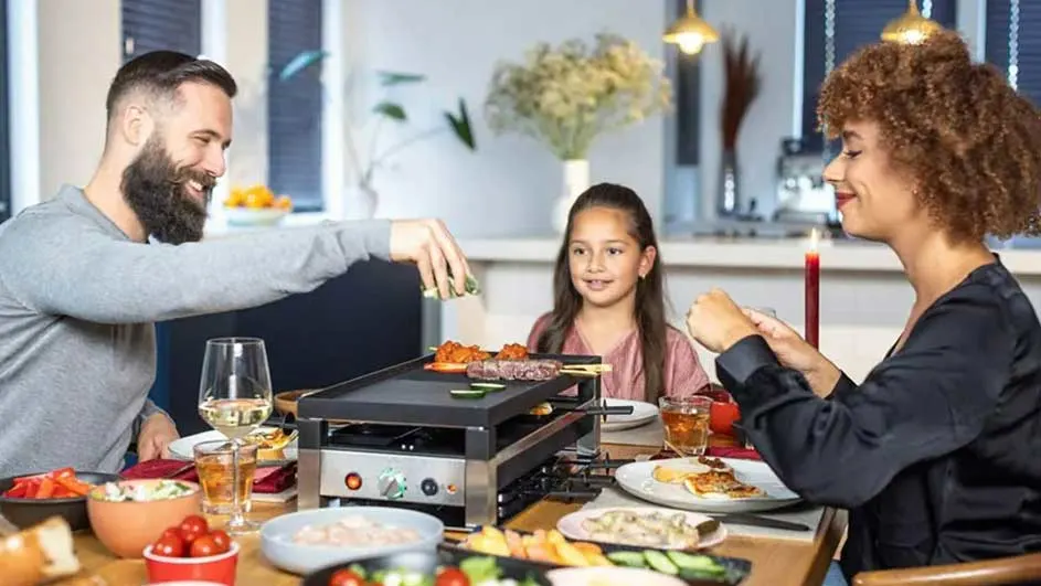 Een tafel met een raclette-grill en ingrediënten. Er staan schalen met groenten en vlees. Achteraan is er een bloemstuk en een raam.