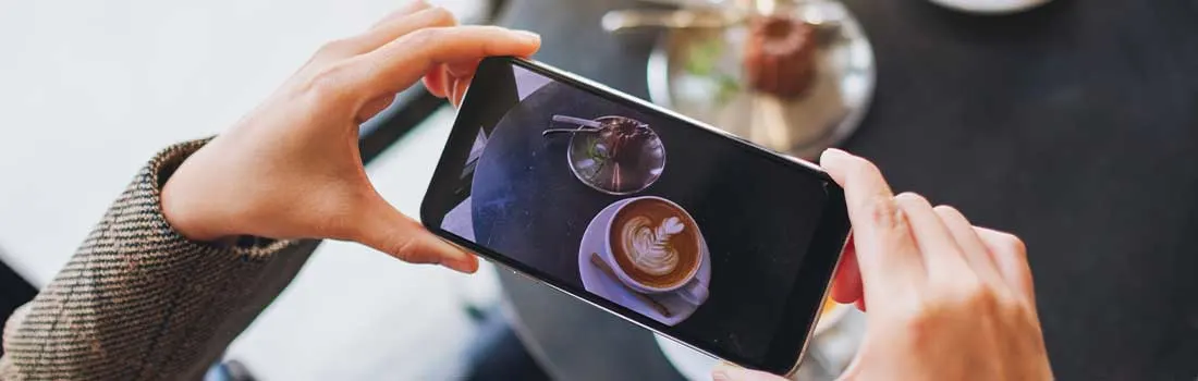Een persoon neemt een foto van een cappuccino en een dessert op een tafel met zijn smartphone, bovenaanzicht. 