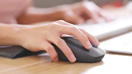 Een hand gebruikt een zwarte, draadloze computermuis op een houten bureau, met een toetsenbord zichtbaar op de achtergrond.