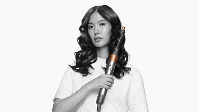 A silver and copper curling iron is held against wavy dark hair, set against a plain white background.