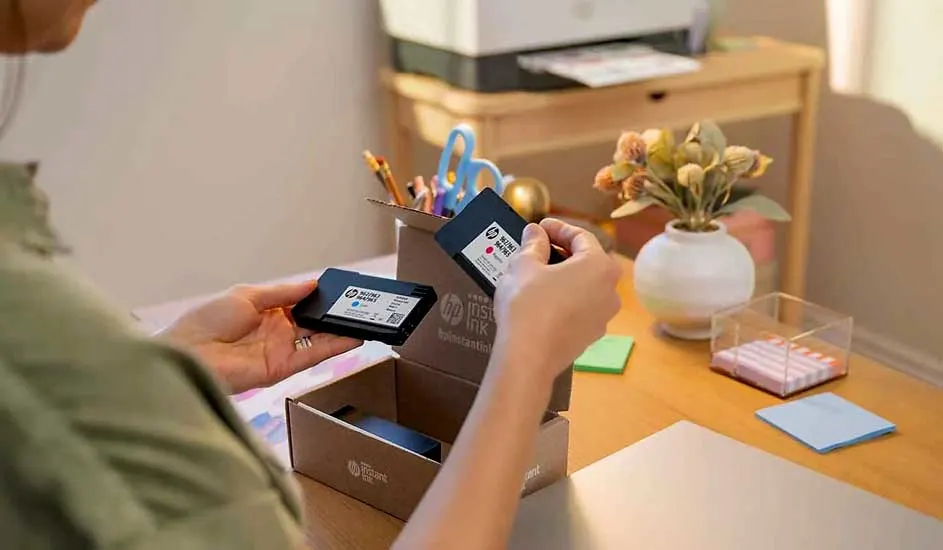 Une personne déballe des cartouches d'encre noires HP sur un bureau en bois avec une imprimante et des fournitures de bureau.
