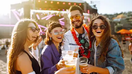 Quatre personnes trinquent avec des verres de bière en plastique à un festival en plein air. Une scène et des lumières sont visibles en arrière-plan.
