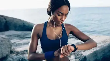 A person in sportswear checks a smart watch. White earphones are visible. The background features rocks and a light blue ocean.