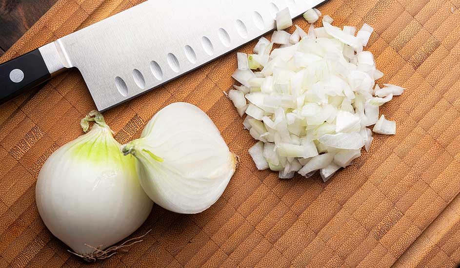 Een houten plank met een zilveren koksmes, twee uien en een hoopje gesneden ui.