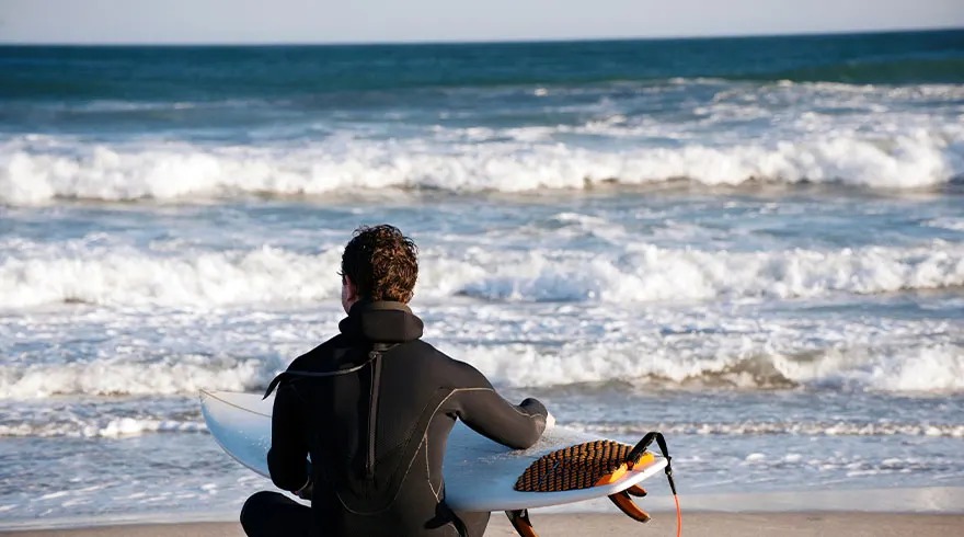 Un surfeur en combinaison noire est assis sur une plage de sable avec une planche blanche, face à l'océan agité.
