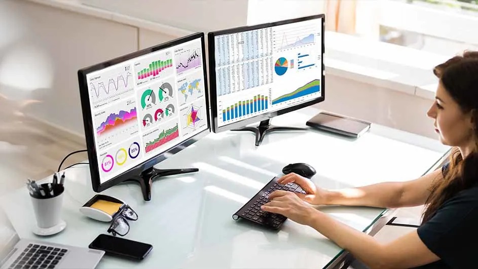 Deux écrans affichent des graphiques colorés sur un bureau blanc avec un clavier noir, une souris, un téléphone et des lunettes.