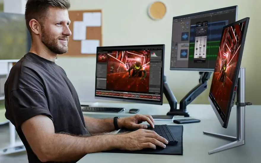 Un bureau avec trois écrans affichant des logiciels de montage vidéo, un clavier et un tapis de souris noirs.