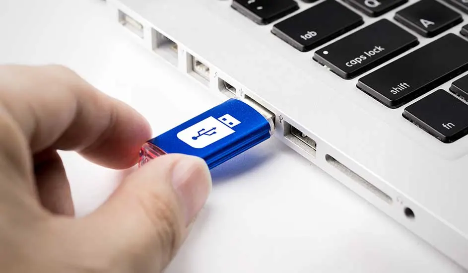Een blauwe USB-stick met een wit USB-symbool wordt in een witte laptop gestoken, gedeeltelijk zichtbaar toetsenbord.