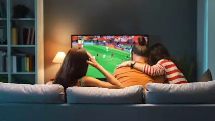 Trois personnes sont assises serrées sur un canapé et regardent un match de football à la télévision dans un salon chaleureux.